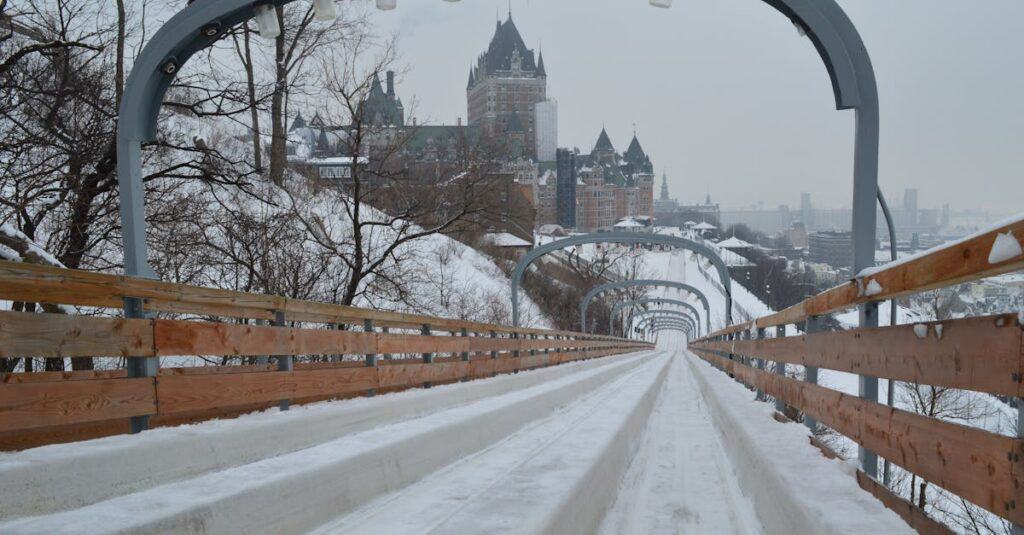 Snow-covered wooden toboggan slide with rails, descending toward a historic castle-like building in a wintery urban landscape.