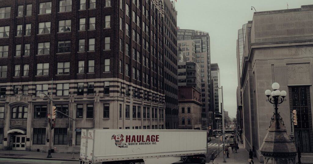 A white Haulage North America Inc. truck drives past tall office buildings on a city street under an overcast sky.