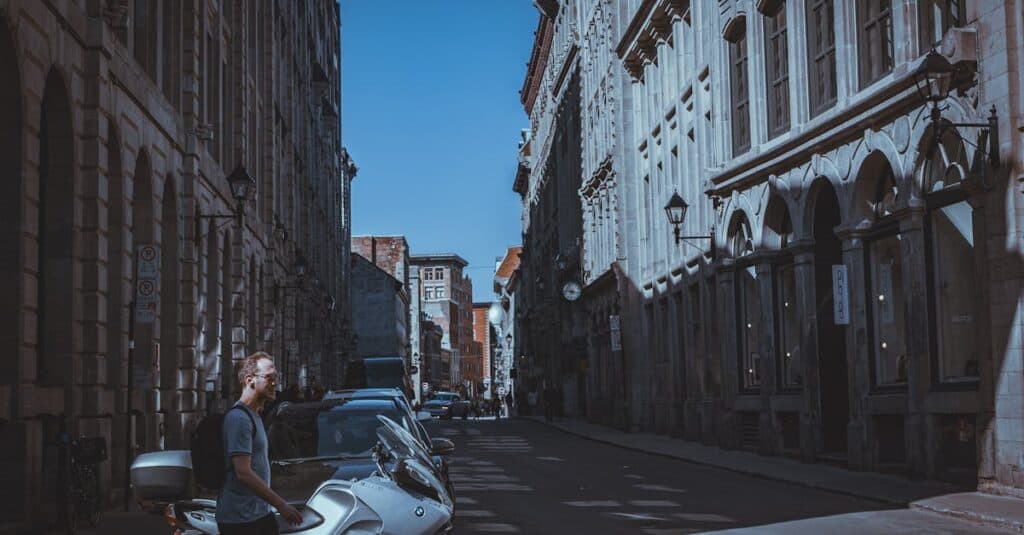 A man stands beside a parked scooter on a narrow, sunlit street lined with old stone buildings and parked cars.