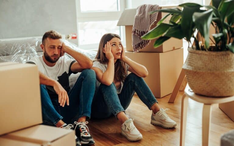 A man and woman sit on the floor surrounded by moving boxes, looking stressed and tired—perhaps wishing they had followed the best checklist for moving houses in Canada. A potted plant and unpacked items are visible in the room.