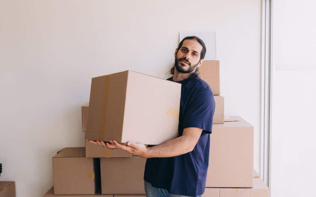 A man with long hair and a beard holds a cardboard box while standing in front of a stack of moving boxes against a white wall.