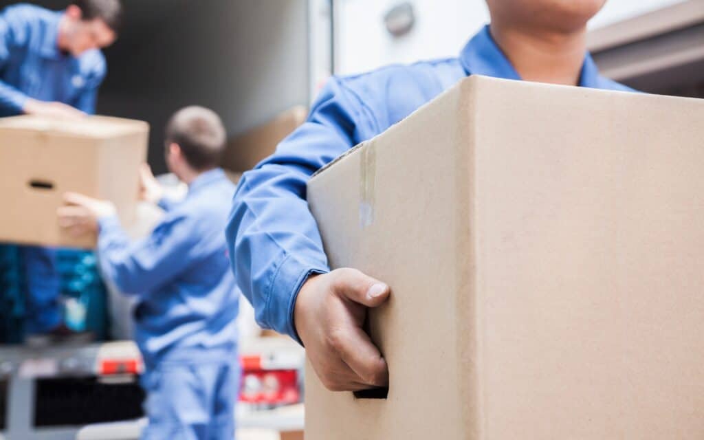 Workers wearing blue uniforms load cardboard boxes into a truck, with one worker in the foreground carrying a box.