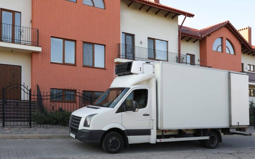 A white refrigerated delivery truck is parked on a street in front of a modern brick and stucco building with balconies.