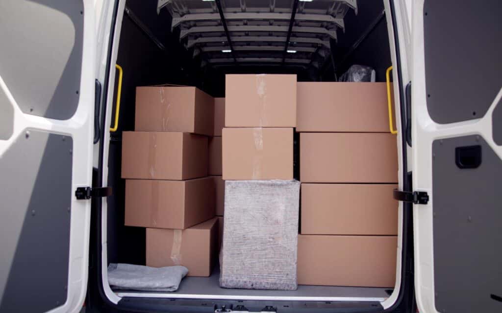 An open van loaded with large cardboard boxes and two wrapped packages, viewed from the back with the doors open.