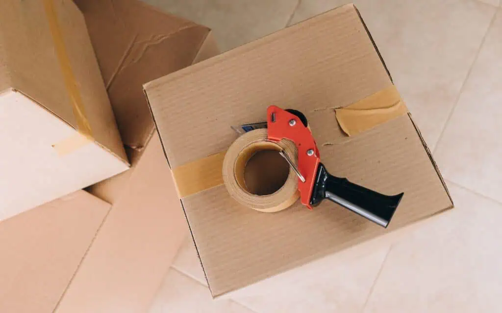 A cardboard box sealed with packing tape sits on a tiled floor, with a tape dispenser placed on top. Other boxes are visible nearby.