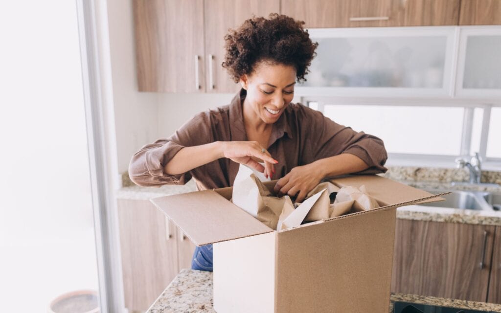 A woman smiles while unpacking or organizing items from a cardboard box on a kitchen counter.
