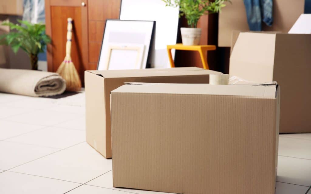 Two cardboard boxes on a tiled floor, with moving supplies, a broom, plants, and rolled carpet visible in the background.