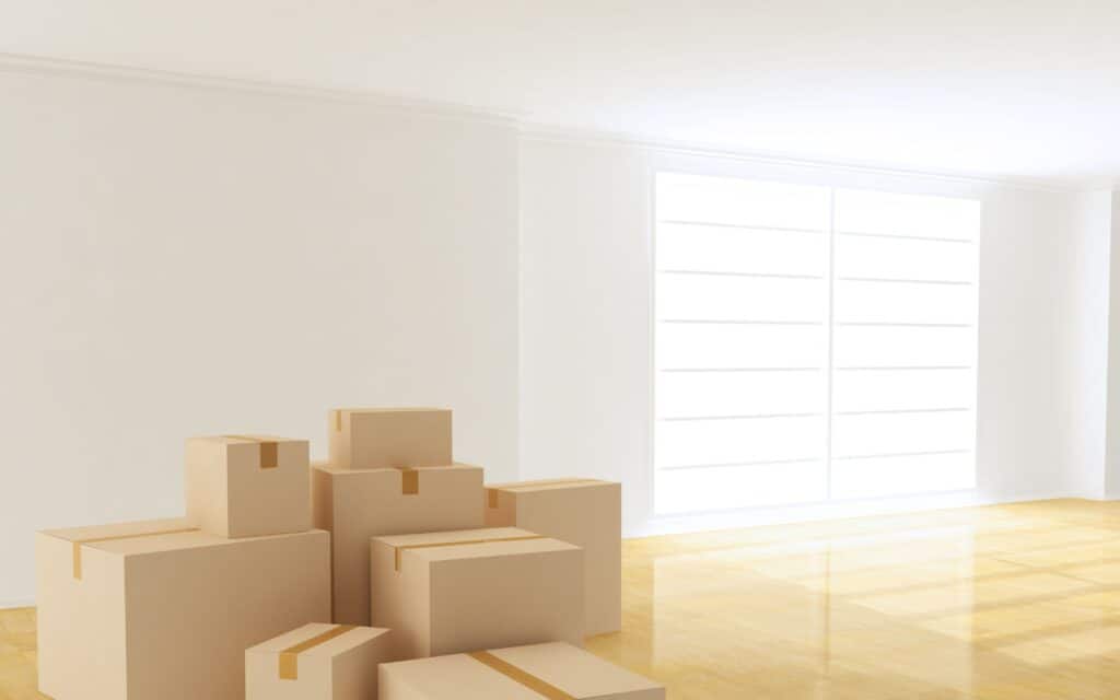 A stack of cardboard boxes sits on a polished wooden floor in an empty, brightly lit room with large windows.