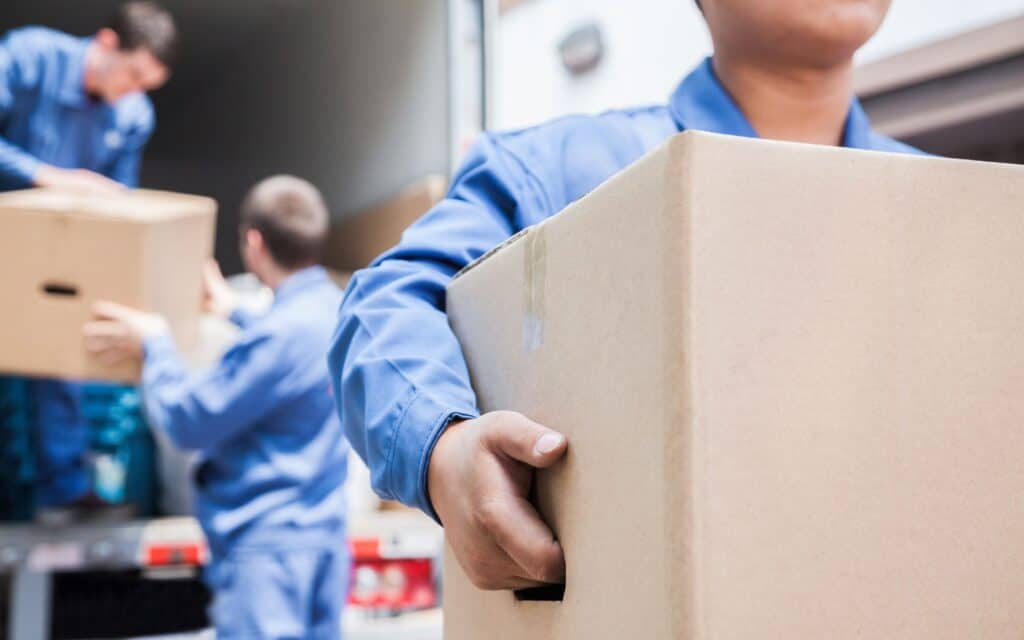 Three people in blue uniforms unload large cardboard boxes from a truck. One person in the foreground is carrying a box.