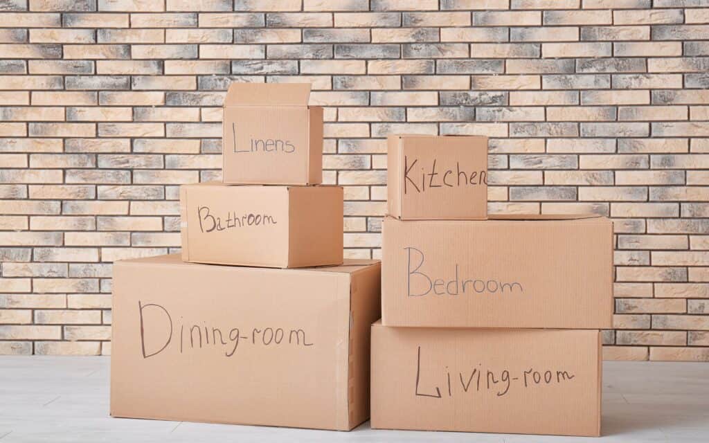 Six labeled moving boxes are stacked against a light brick wall. Labels read: Dining-room, Bathroom, Linens, Kitchen, Bedroom, and Living-room.