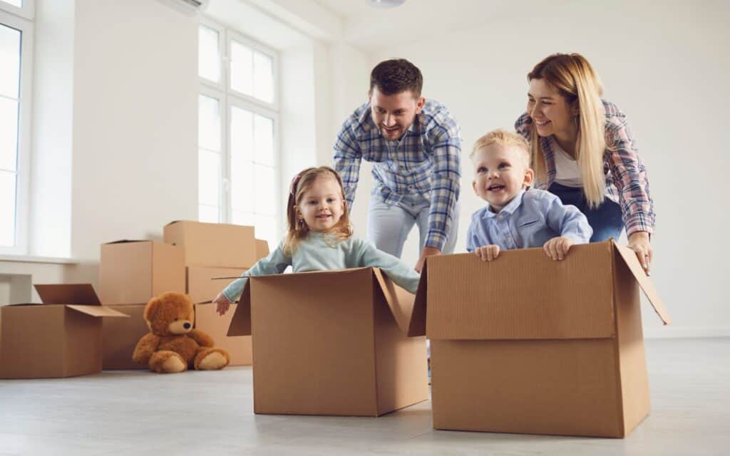 Two adults push two young children who are sitting in cardboard boxes in a room with unpacked boxes and a teddy bear.