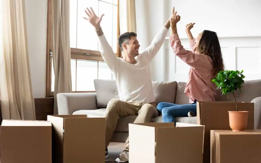 A man and woman sit on a couch surrounded by moving boxes, raising their arms and smiling, appearing to celebrate.