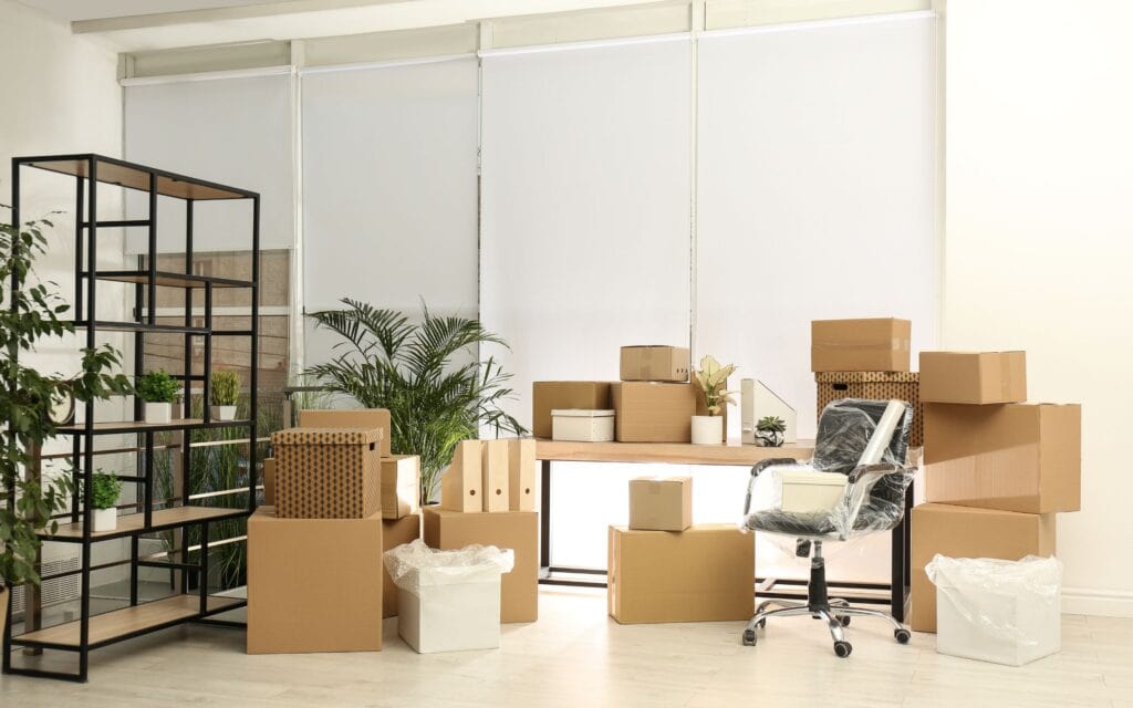 Office space with desk, chair, shelves, and several cardboard boxes and plants, suggesting a move or relocation.