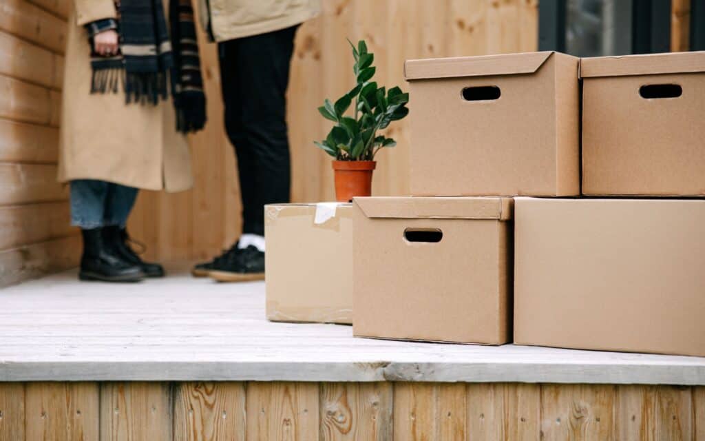 Several cardboard boxes and a potted plant are stacked on a wooden porch. Two people stand in the background, partially visible.