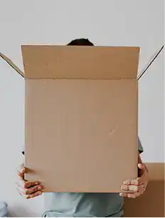 A person holds a large, open cardboard box in front of their face, obscuring their head and upper body.