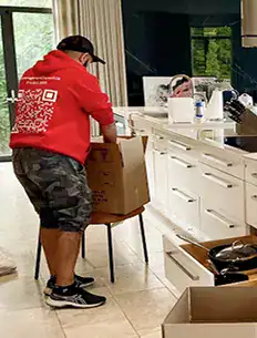 A person in a red hoodie and camo shorts packs items into a cardboard box in a modern kitchen.