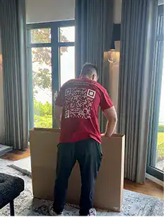 A person wearing a red shirt with a large QR code on the back stands in front of a cardboard box inside a room with tall windows and curtains.