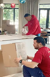 Two men in red shirts pack items into cardboard boxes in a modern, well-lit room with large windows and hanging light fixtures.